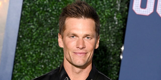 Tom Brady in a white shirt and leather jacket smiles gently and looks directly into the camera in the "80 for Brady" Red carpet