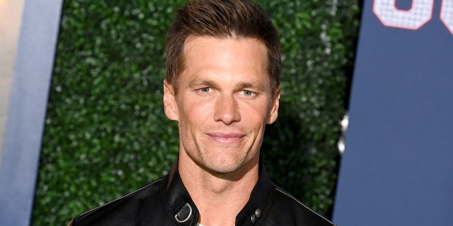 Tom Brady in a white shirt and leather jacket smiles gently and looks directly into the camera in the "80 for Brady" Red carpet