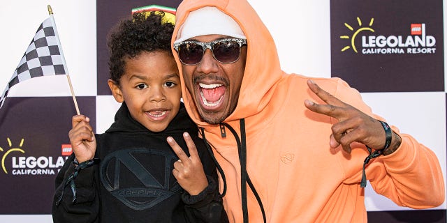 Nick Cannon in an orange sweatshirt, white band around his head and dark sunglasses poses with his son Golden Cannon doing a peace sign while wearing black on the red carpet