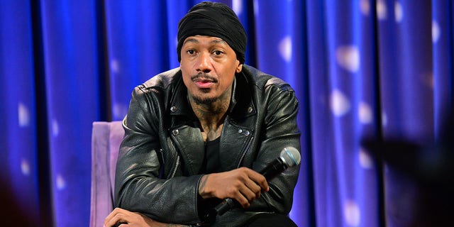 Nick Cannon with a black turban leans over in a black leather jacket sitting on a purple chair holding a microphone