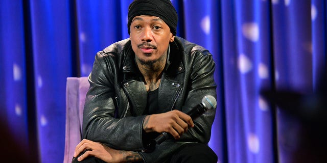 Nick Cannon with a black turban leans over in a black leather jacket sitting on a purple chair holding a microphone