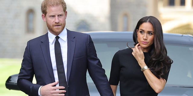 Prince Harry in a navy suit and tie holds Meghan Markles hand, wearing a dark dress after the Queen passed away