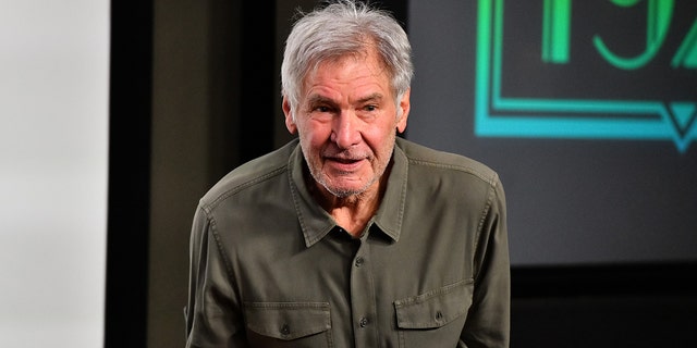 Harrison Ford in an olive green shirt with pockets speaks to a crowd