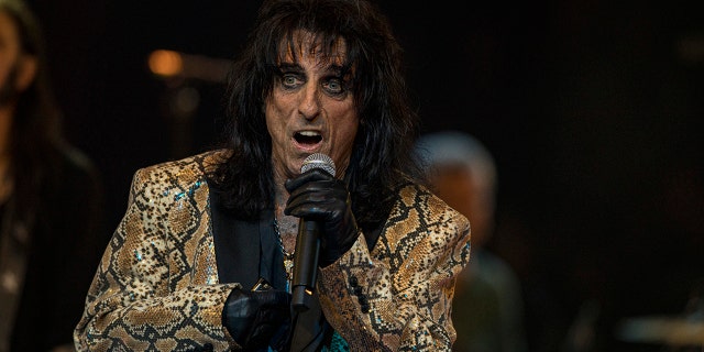 Jeff Beck in a gold and black snakeskin jacket and a thick layer of eyeliner holds up a microphone on stage