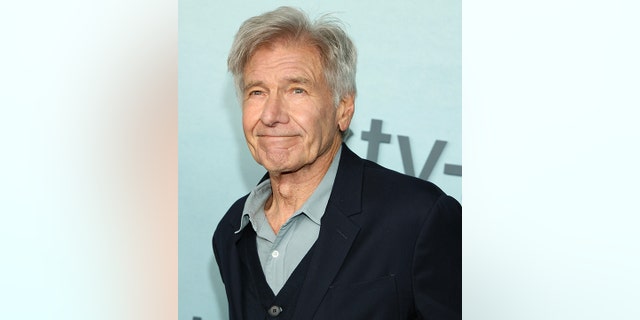 Harrison Ford soft smiles in a light turquoise shirt and black suit jacket on the red carpet