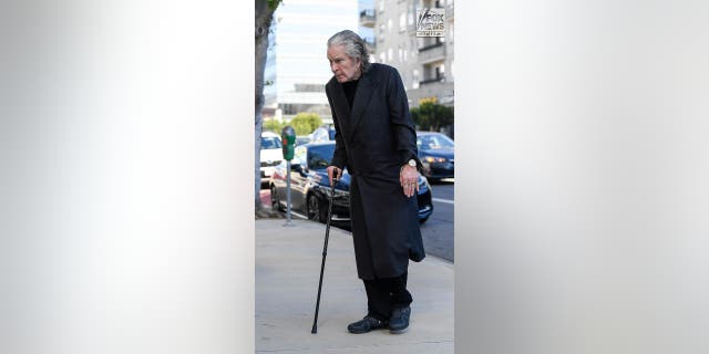 Ozzy Osbourne wearing a black coat over dark clothes walks with a cane