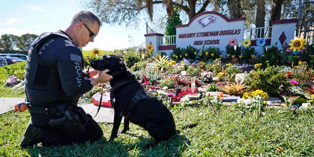 Coconut Creek, Fla., police officer Michael Leonard takes a moment with his School Safety Dog Taylor, Tuesday, Feb. 14, 2023, as he visits a memorial in front of Marjory Stoneman Douglas High School (MSD) in Parkland, Fla. Leonard was the officer that stopped and arrested the shooter Nikolas Cruz.