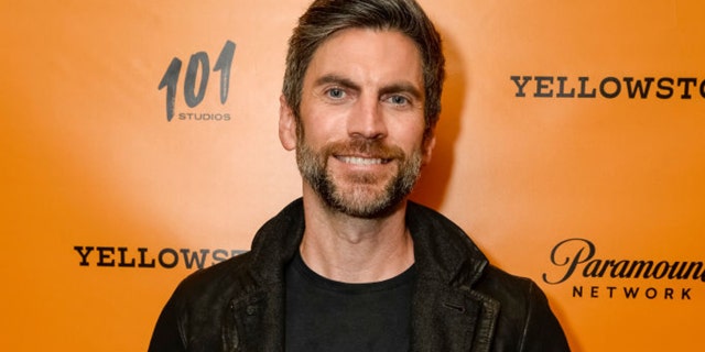 Wes Bentley in black smiles in front of an orange backdrop for a "Yellowstone" premiere