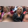 A woman wearing a black jacket listens from the gallery inside a courtroom.
