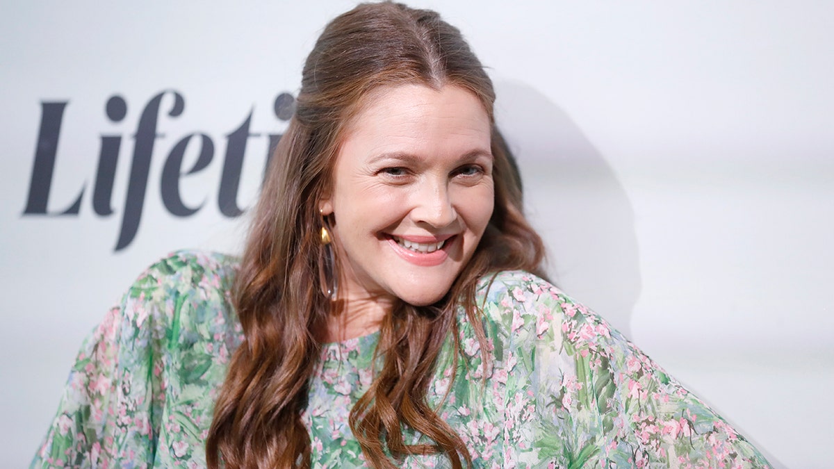 Drew Barrymore smiles for a photo with her shoulders slightly shrugged in a green and pink dress