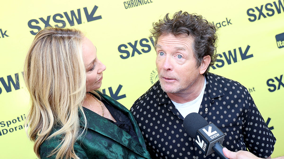 Tracy Pollan in a green jacket looks lovingly at Michael J. Fox as he speaks into a microphone on the SXSW carpet wearing a black shirt with white flowers on it