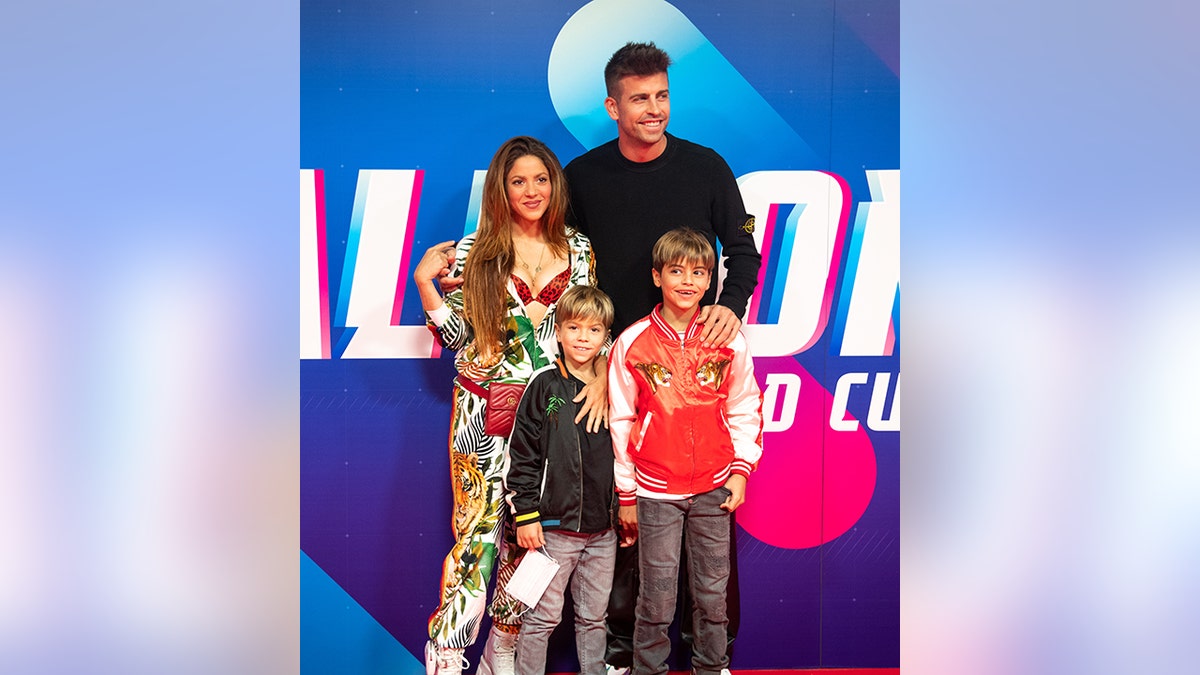 Shakira exposes her red leopard bra wearing a white outfit with jungle animals on it, has Gerard Piqué's arm wrapped around her, he is wearing black, and their two sons both in relaxed bomber jackets stand in front of them