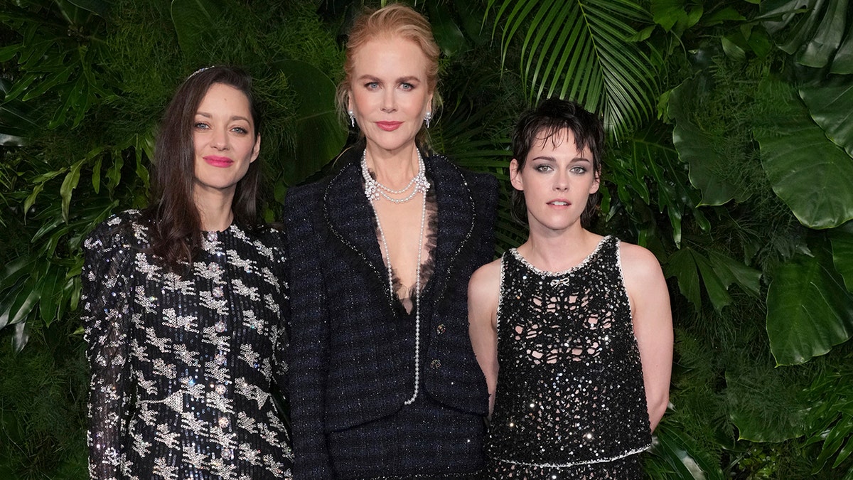 Marion Cotillard in a sparkly black jacket stands next to Nicole Kidman in a navy and black tweed jacket and skirt set next to Kristen Stewart in a see-through black dress with sparkles