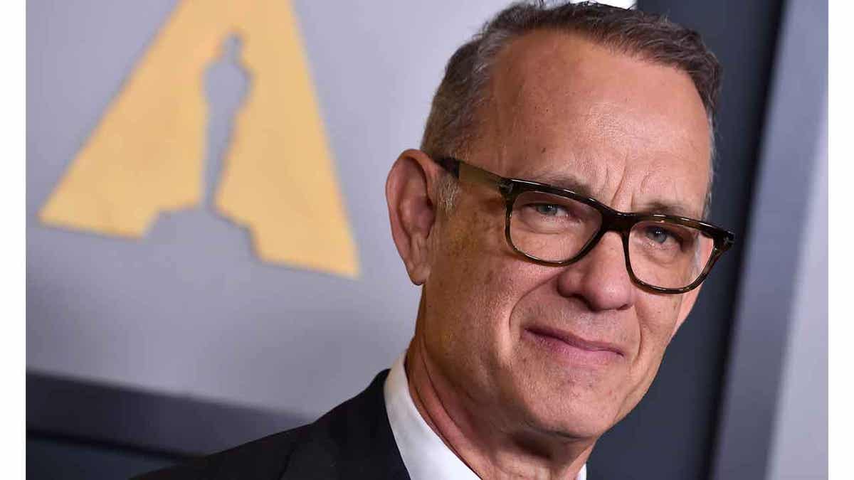 Tom Hanks soft smiles on the red carpet in a dark suit and dark framed glasses