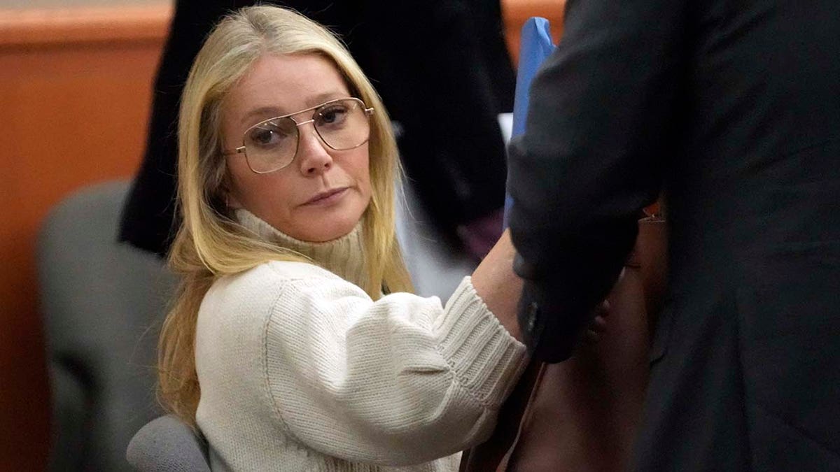 Gwyneth Paltrow wears beige sweater and retro glasses and sits next to lawyers in court