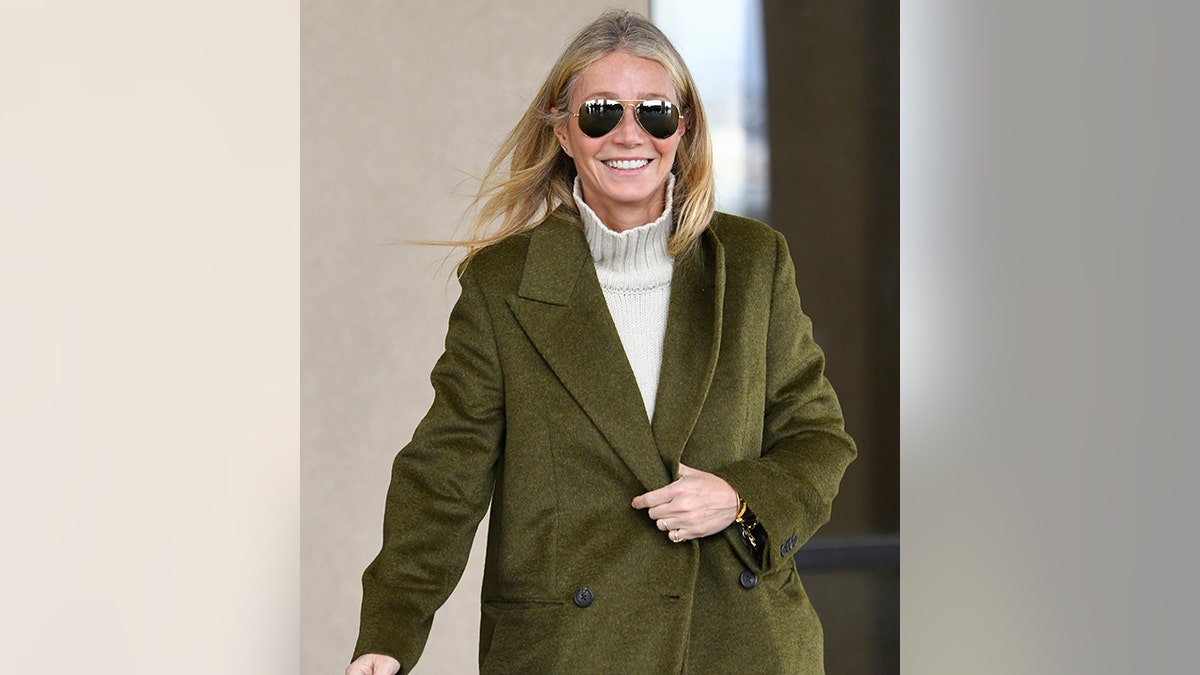 Gwyneth Paltrow smiles in a long green jacket and light turtleneck behind a pair of sunglasses as she leaves court