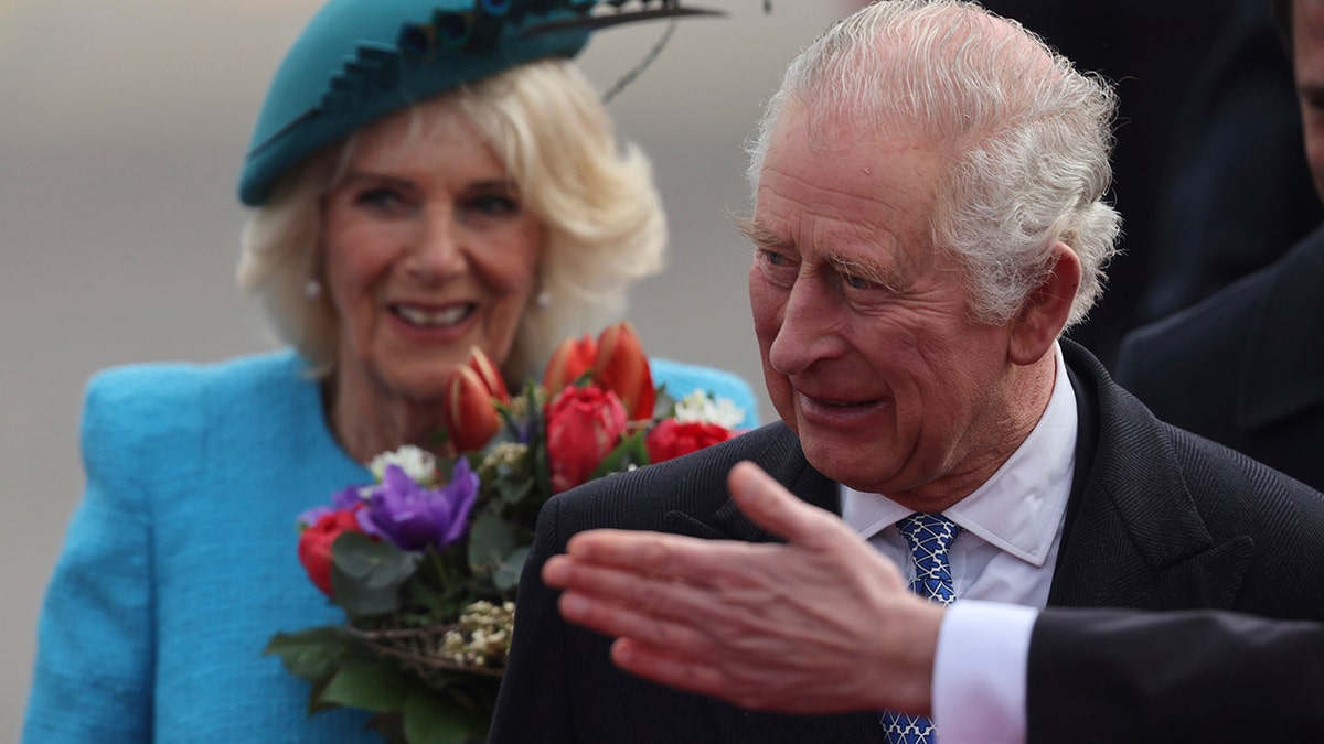 King Charles looks off to supporters as he arrives in Germany and a hand ushers him to the right, Camilla in a turquoise/teal outfit walks behind with a bouquet of flowers