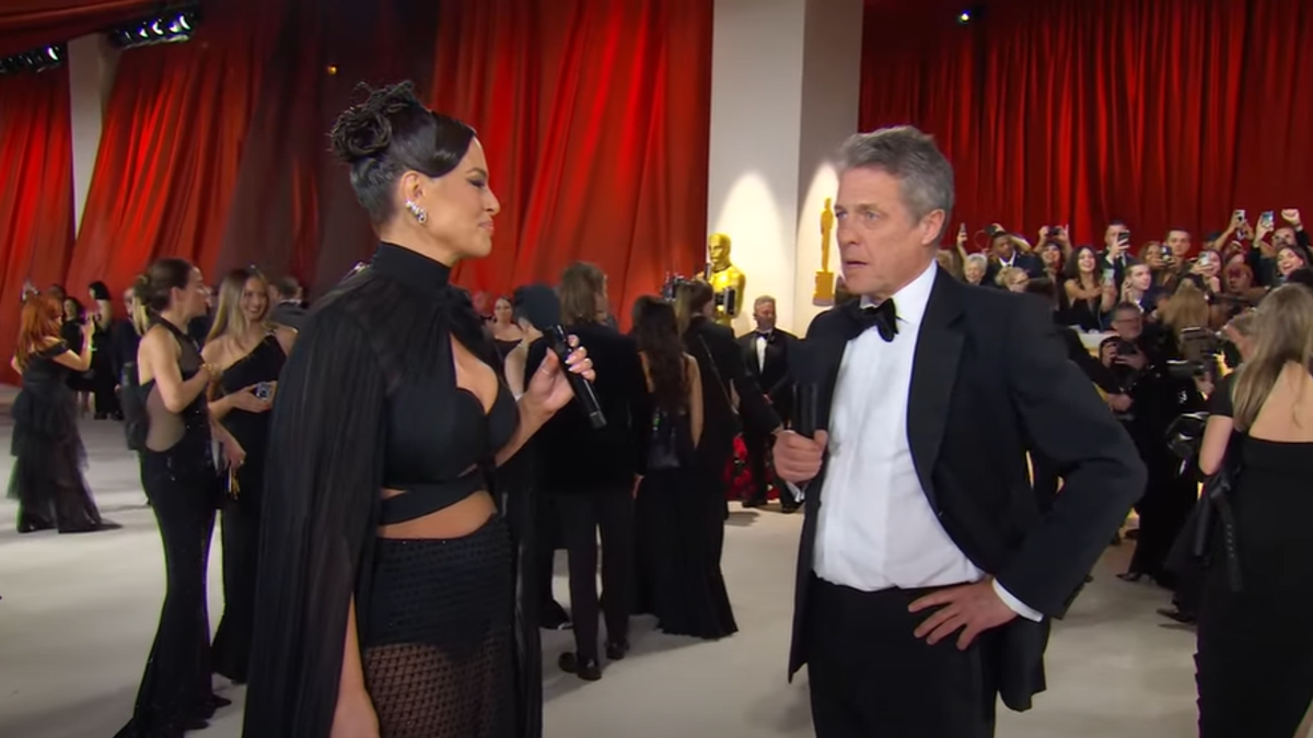 Ashley Graham holds a microphone and interviews Hugh Grant