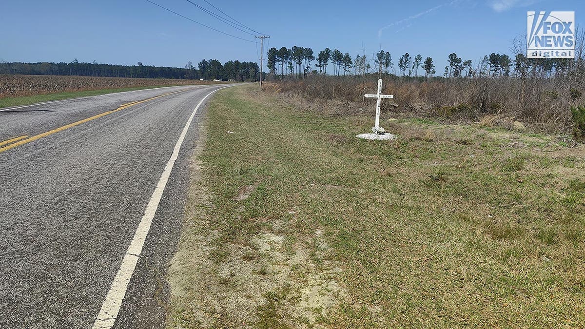 A white wooden cross with Stephen Smith's name and lifespan dates on it is on a grassy area by the side of the road