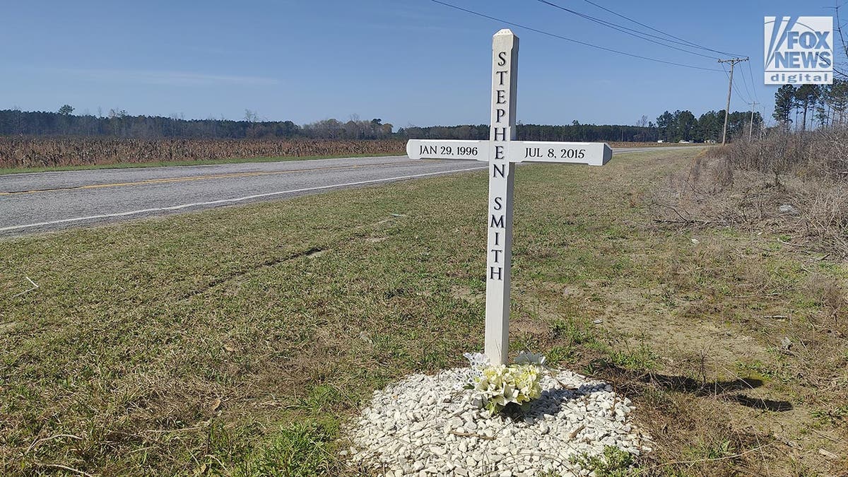 A white wooden cross with Stephen Smith's name and lifespan dates on it is on a grassy area by the side of the road