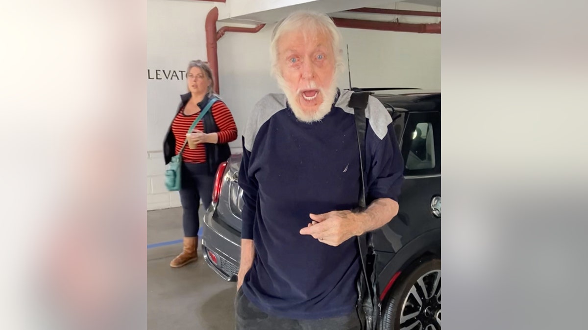 Dick Van Dyke in a navy shirt looks directly at the camera in a parking garage with his wife Arlene behind him wearing a red shirt