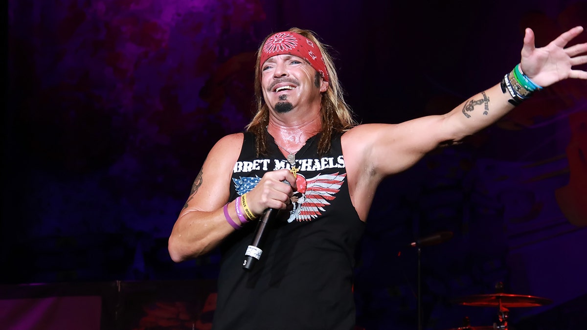 Bret Michaels on stage in a black tank top that says his name on it, red bandana, with a microphone in his hand and arm extended