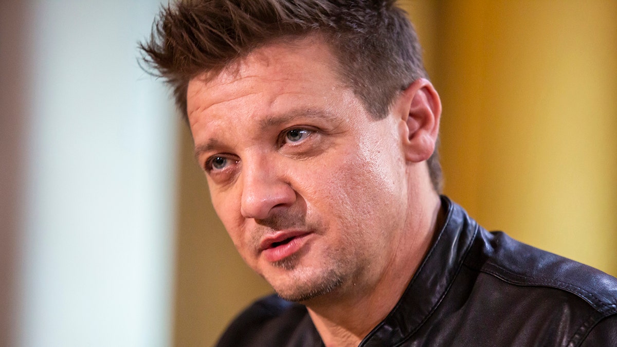 Jeremy Renner wearing a black leather jacket looks serious as he speaks to Willie Geist (off-camera)