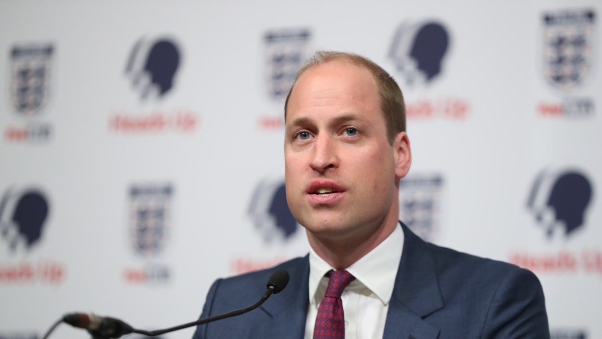 Prince William in a blue suit and red tie speaking at a small microphone