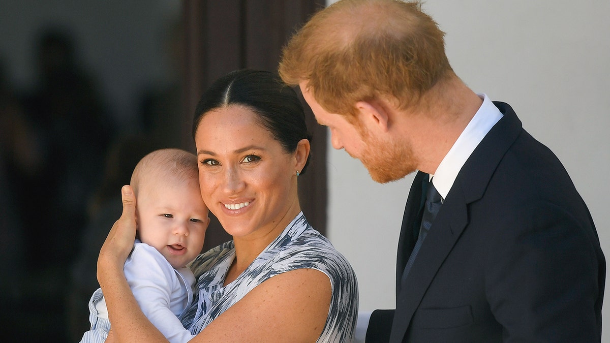 Meghan Markle in a printed dress holds Prince Archie as an infant as Prince Harry looks at them while in South Africa