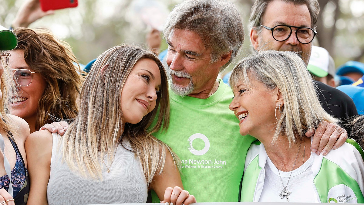 Chloe Lattanzi in a white shirt stares at her mother Oliva Newton-John lovingly also in a white shirt and jacket with green trim, in between them is John Easterling, wearing a green t-shirt at Olivia's Walk for Wellness event in Australia