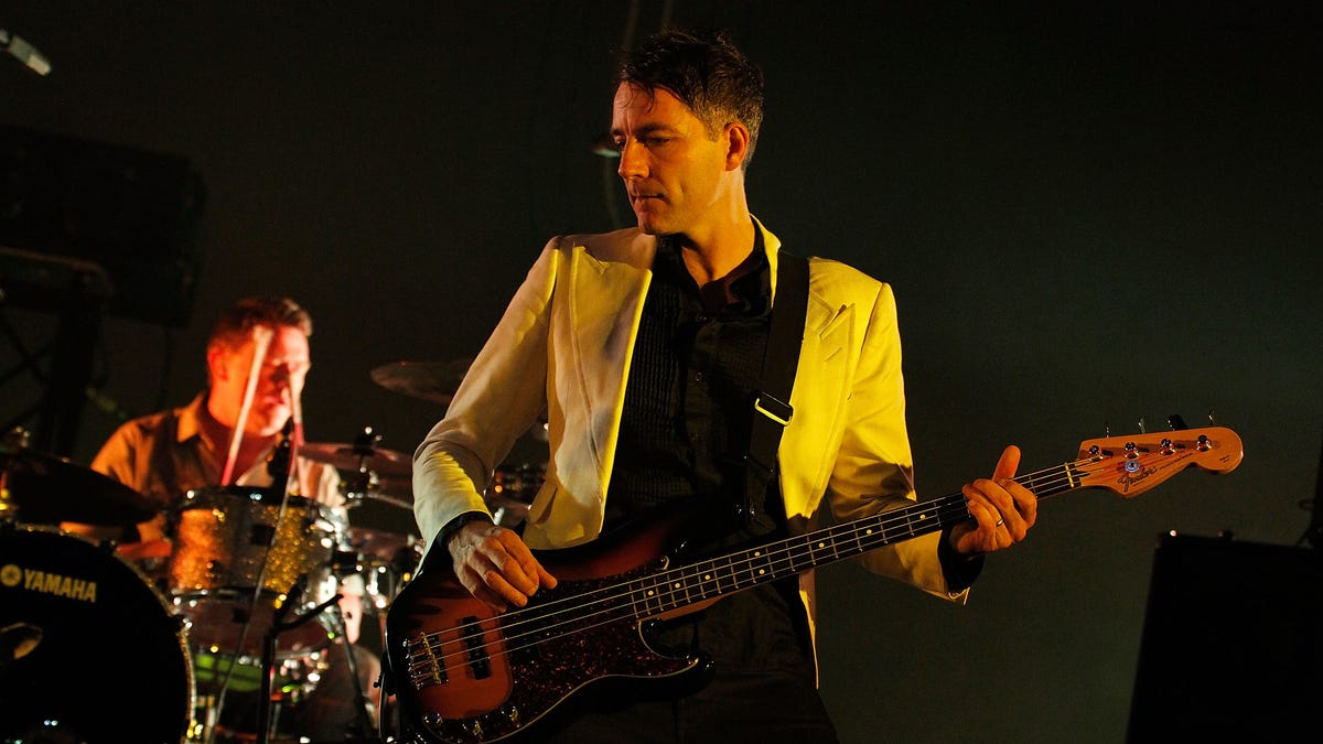 Steve Mackey wears a white jacket and plays bass guitar