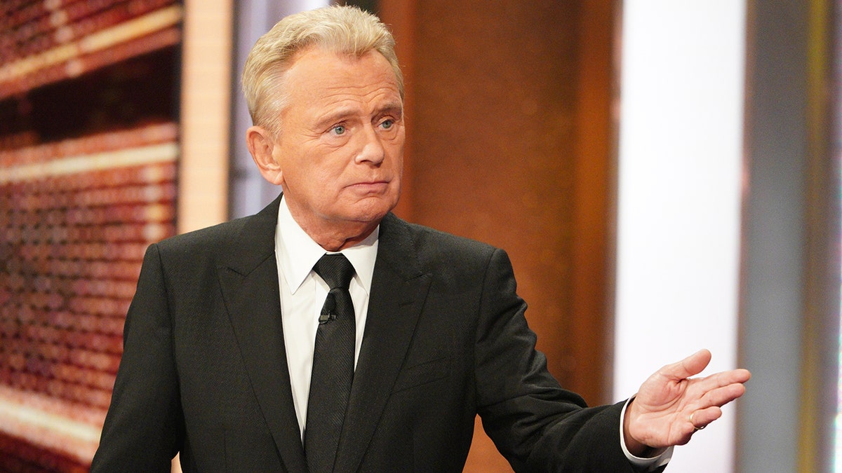 Pat Sajak in a black suit, white button down, and black tie looks off-camera while filming "Celebrity Wheel of Fortune"
