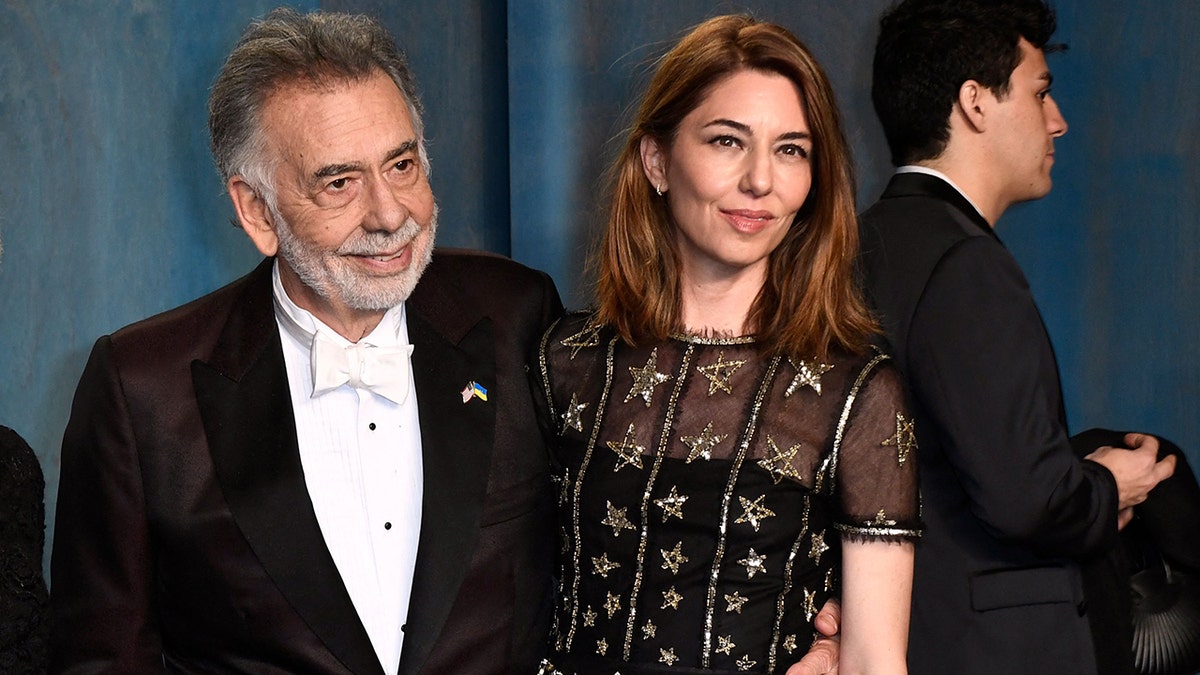 Francis Ford Coppola in a black tuxedo, with a white shirt and white bow-tie smiles next to daughter Sofia in a black dress with sequined gold stars embroidered and a mesh top at the Vanity Fair Oscar party