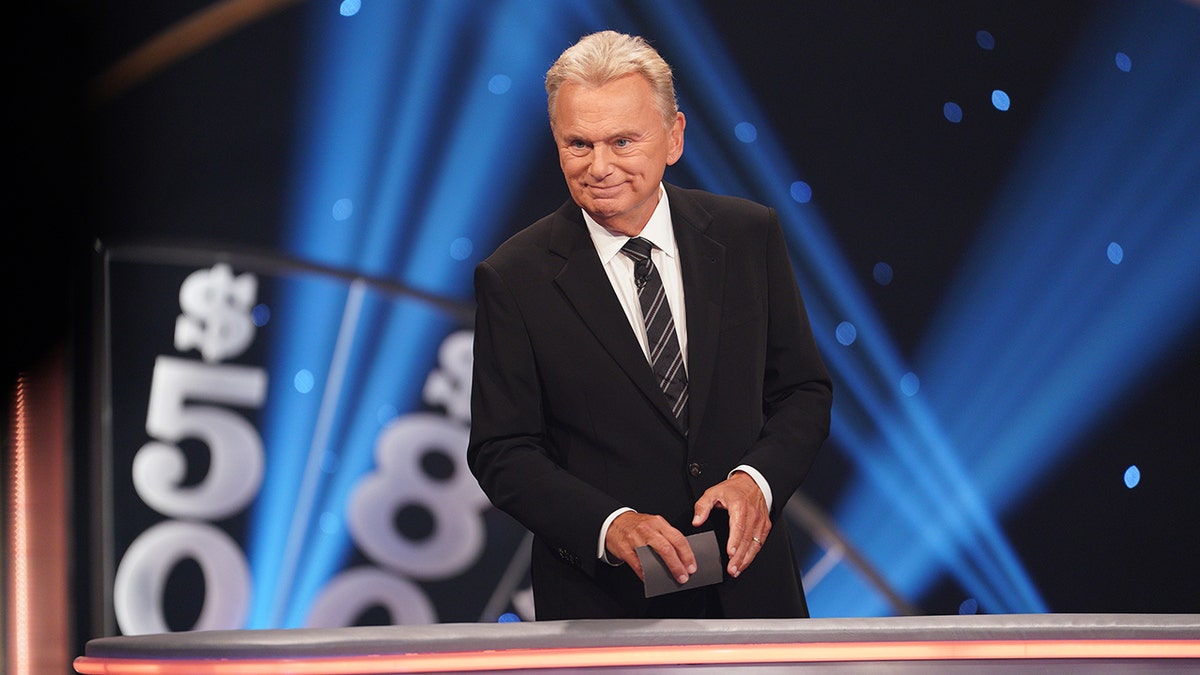 Pat Sajak stands behinds a barrier wearing a black suit and a patterned tie holding a card taping "Celebrity Wheel of Fortune"