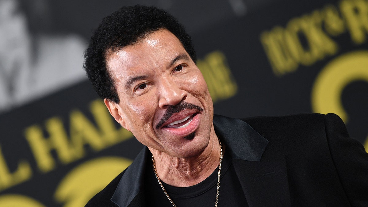 Lionel Richie smiles with a gold chain around his neck, velvet black suit and black shirt