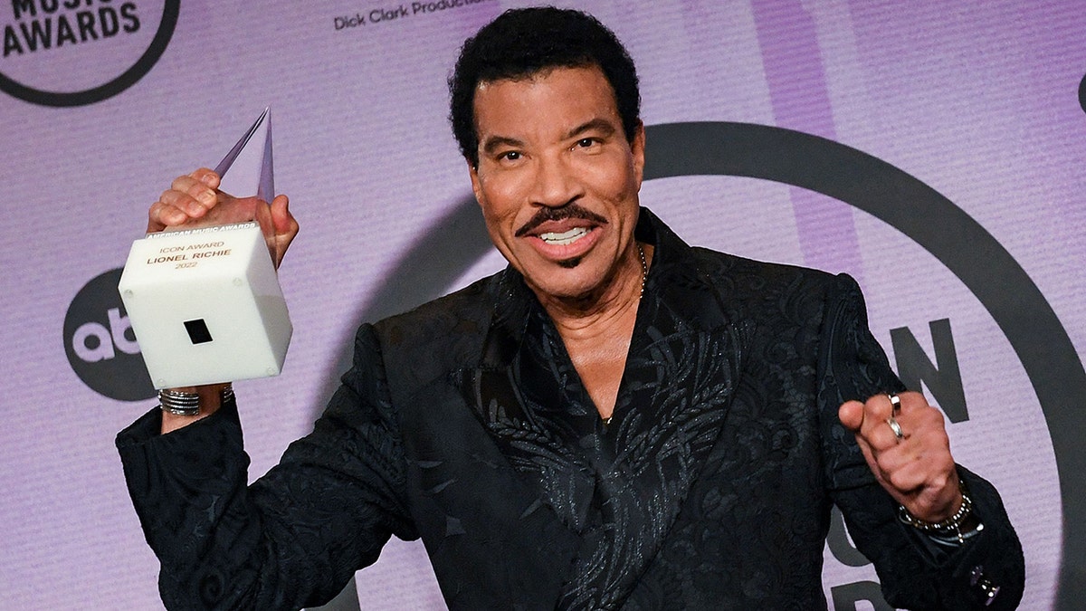 Lionel Richie in a black patterned jacket holds up an award on the red carpet at the American Music Awards