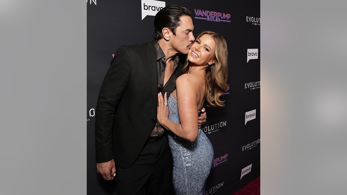 Tom Sandoval in a black suit kisses girlfriend Ariana Madix in a sparkly blue dress, as she lays her hand on his chest