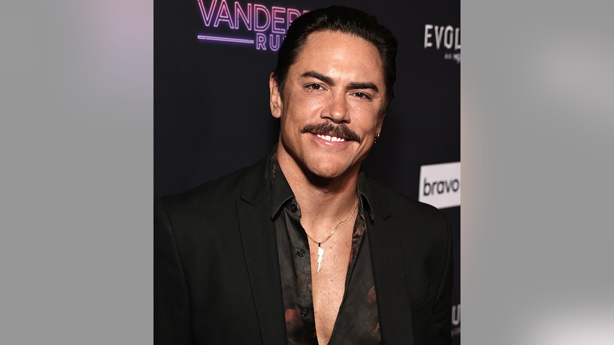 Tom Sandoval wears a black printed top and black suit and smiles for a photo with a dark mustache