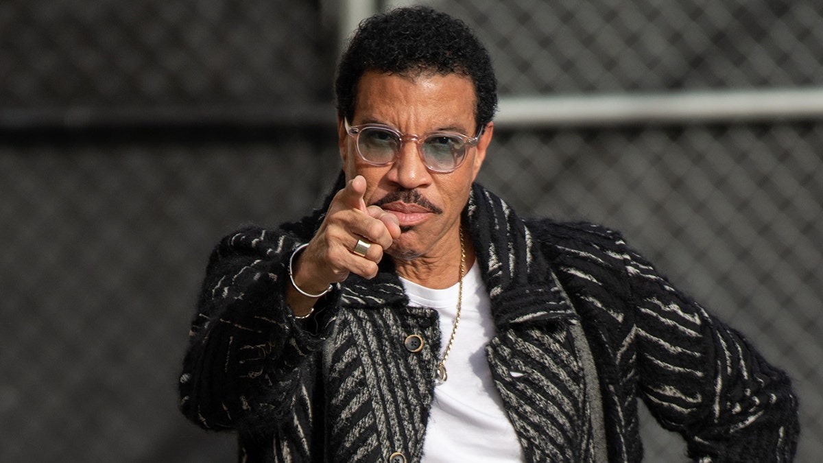 Lionel Richie in a striped black jacket, white t-shirt and chain with blue-lens glasses points at the camera out in Los Angeles