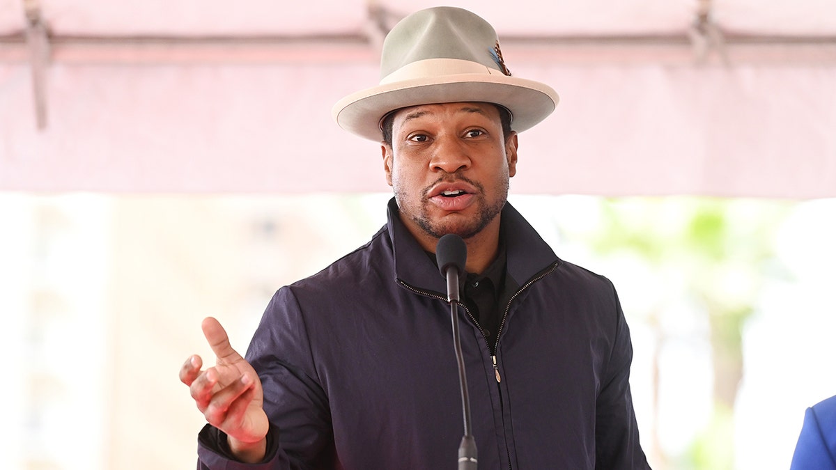 Jonathan Majors speaks at the podium for actor Michael B. Jordan's Star unveiling on the Hollywood Walk of Fame, wearing a blue jacket and large tan hat