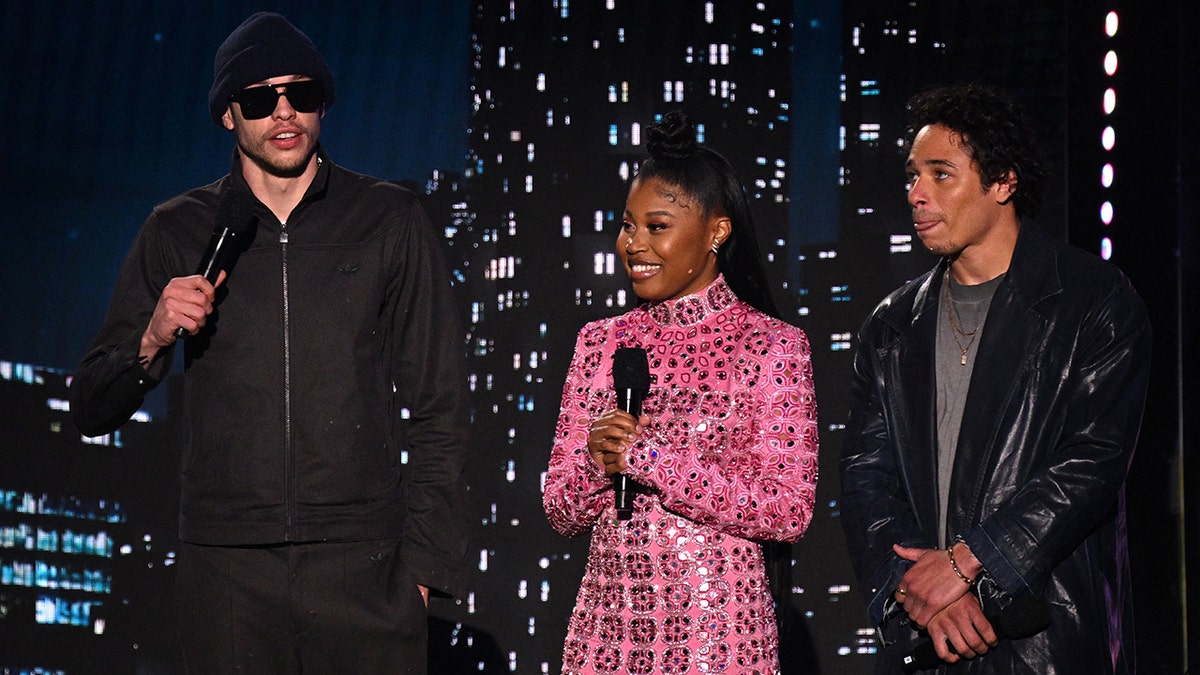 Pete Davidson in all-black, wearing sunglasses and a black hat next to Dominique Fishback in a pink sparkly turtleneck dress and Anthony Ramos in a black leather jacket presenting on stage at the Kids' Choice Awards