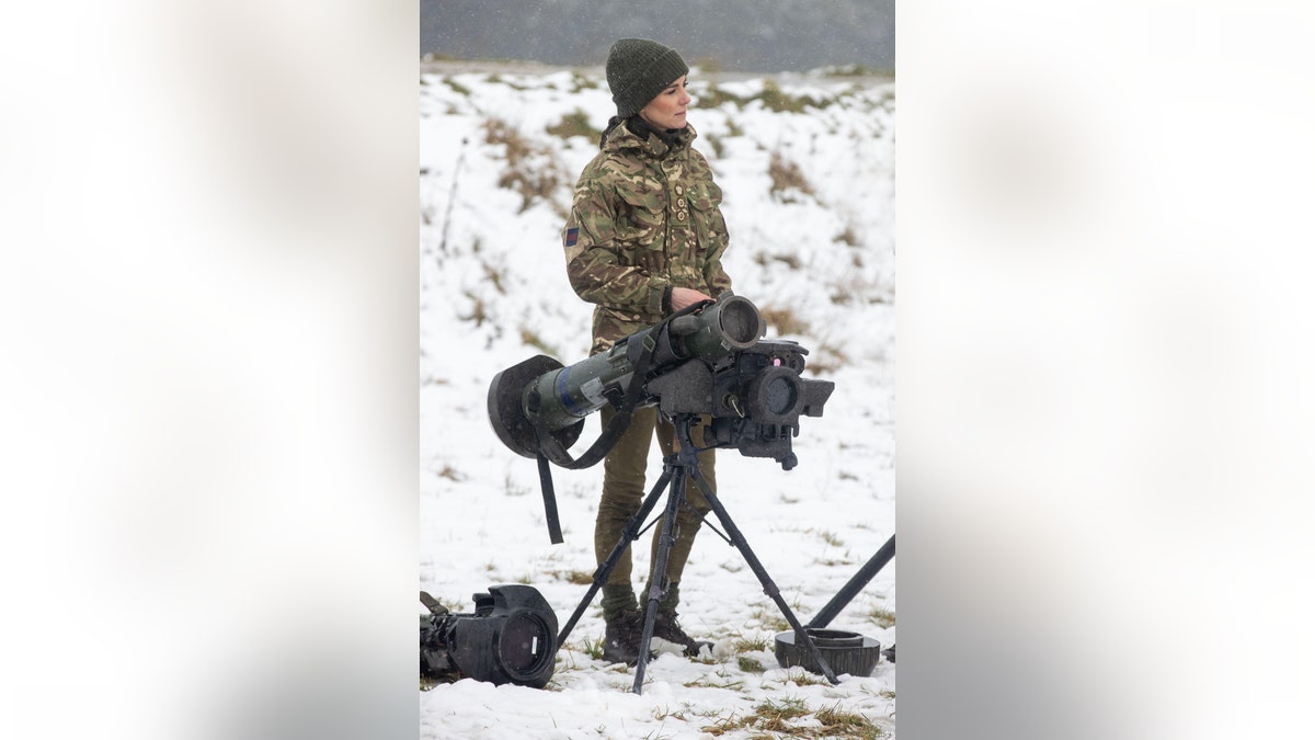 Kate Middleton in camo stands in snowy field next to military weapon