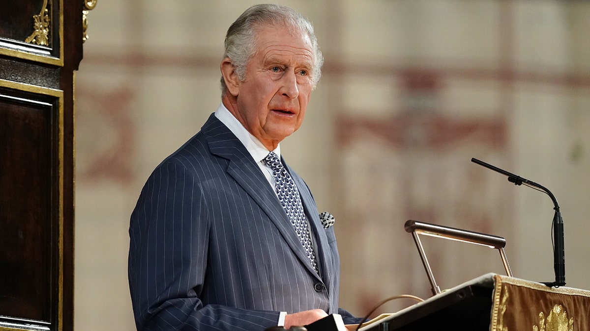 King Charles at the podium wearing a blue pin-stripe suit speaking in Westminster Abbey for Commonwealth Day