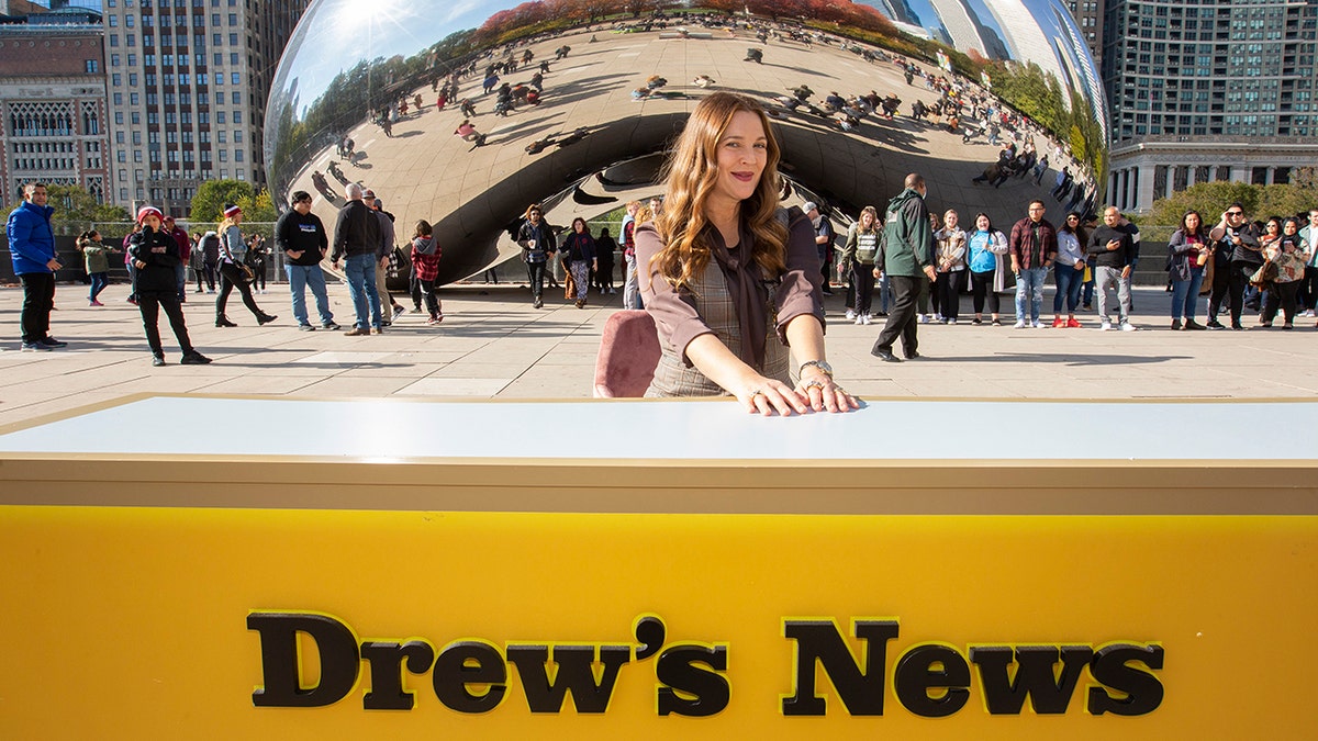 Drew Barrymore in a brown shirt and plaid vest smiles in front of the "Bean" statue in Chicago while sitting behind a desk that reads "Drew's News," taping a segment for her talk show