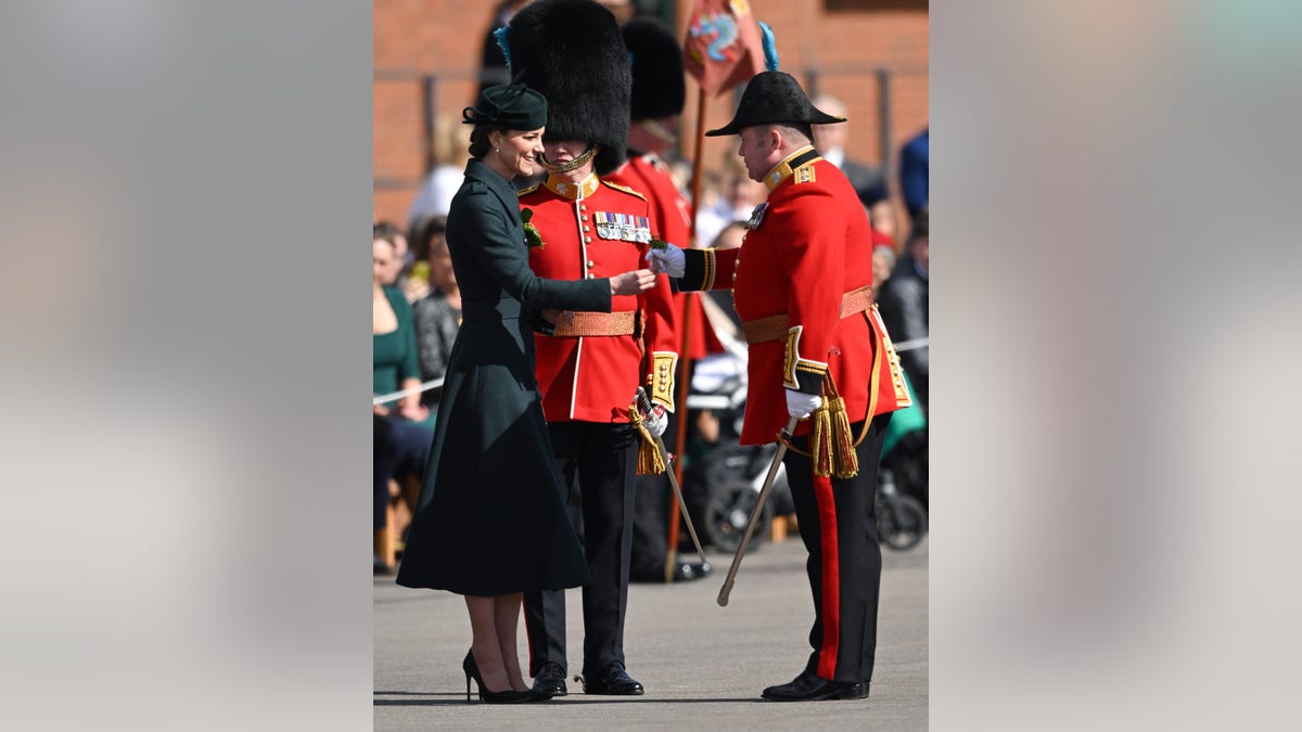 Kate Middleton in green jacket dress and black hat hands shamrocks to Irish Guard in red uniform