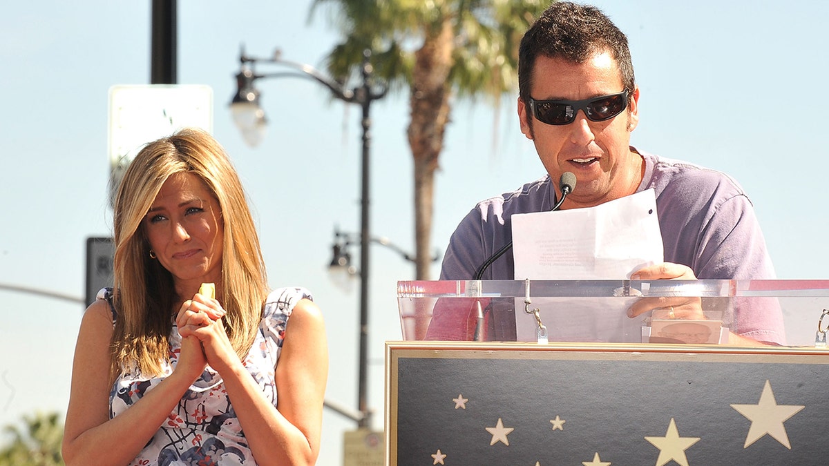 Adam Sandler giving a speech at Jennifer Aniston's Walk of Fame ceremony, with her standing behind him.