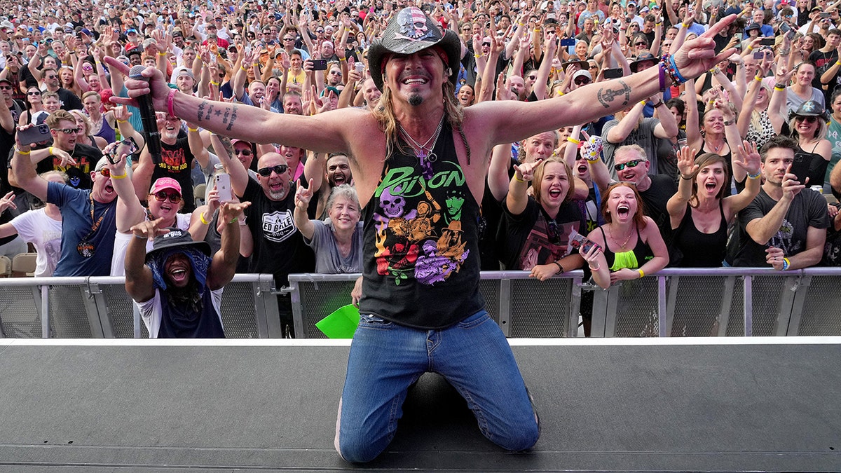 Bret Michaels in a black tank top, jeans, on his knees, back to the crowd with a black cowboy hat and arms spread out during a concert