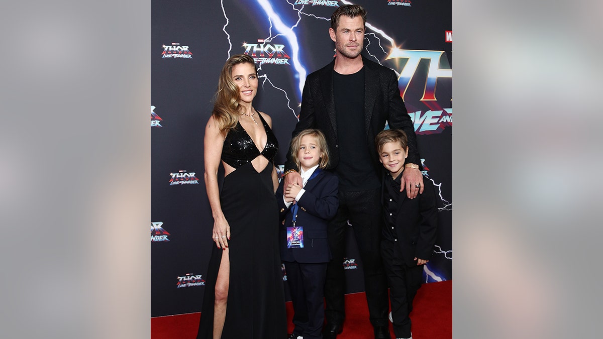Chris Hemsworth de camisa e jaqueta pretas sorri com seus dois filhos (um de terno preto completo, o outro de terno preto e camisa branca) junto com sua esposa Elsa Pataky em um vestido preto recortado no tapete vermelho