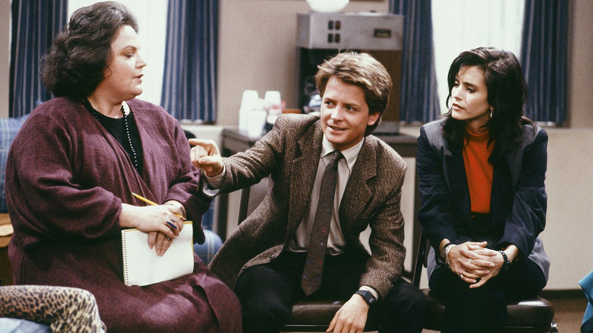 Michael J. Fox in a brown suit and tie sits on a chair as Alex P. Keaton in "Family Ties" alongside Courteney Cox who played his girlfriend Lauren Miller in a red top and black suit, speaking to Diana Bellamy as Dr. Eileen Davidson in a dark purple sweater