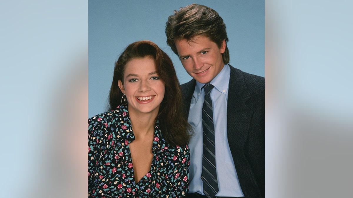 Justine Bateman as Mallory Keaton in a patterned blouse smiles for a photo with Michael J. Fox as Alex Keaton in suit, light blue shirt, and dark blue tie for "Family Ties"